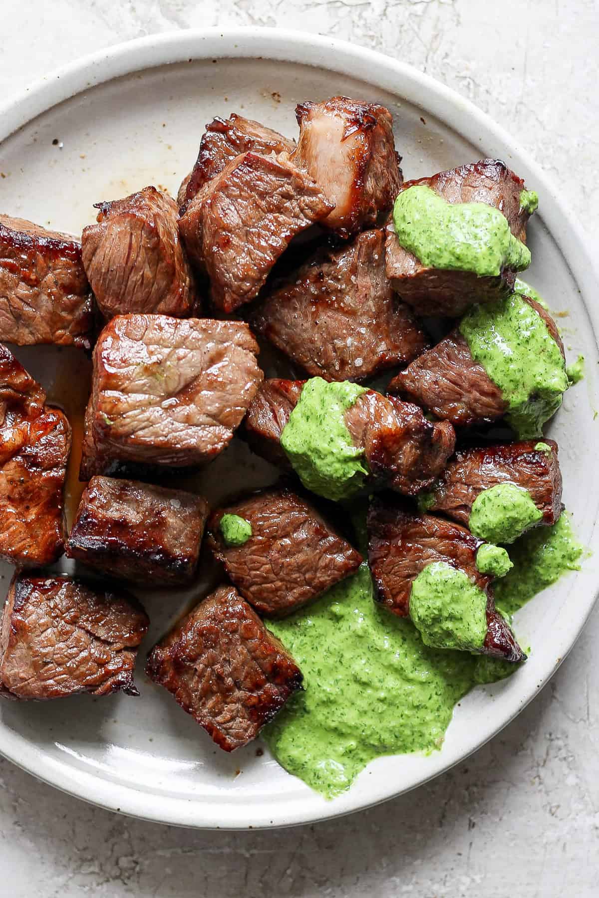 A white plate with grilled steak bites topped with a green herb sauce.