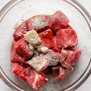 A glass bowl filled with raw beef chunks seasoned with spices on top, placed on a light-colored textured surface.