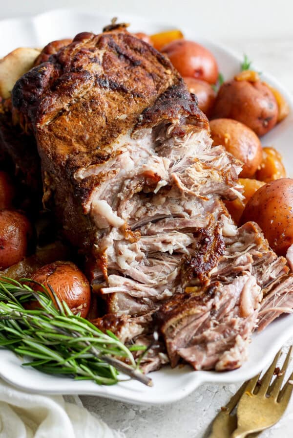 A close-up of a seasoned, cooked roast surrounded by whole red potatoes and fresh rosemary sprigs on a white platter. A fork and knife are set to the side.