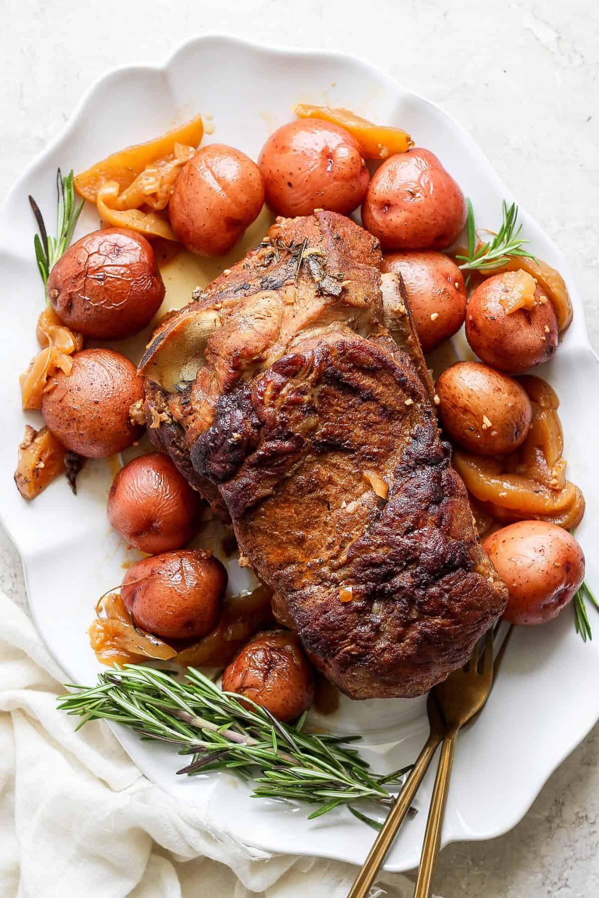 A roasted pork shoulder is served on a white plate, accompanied by red potatoes, onions, and garnished with rosemary sprigs. A white cloth is partially visible on the side.