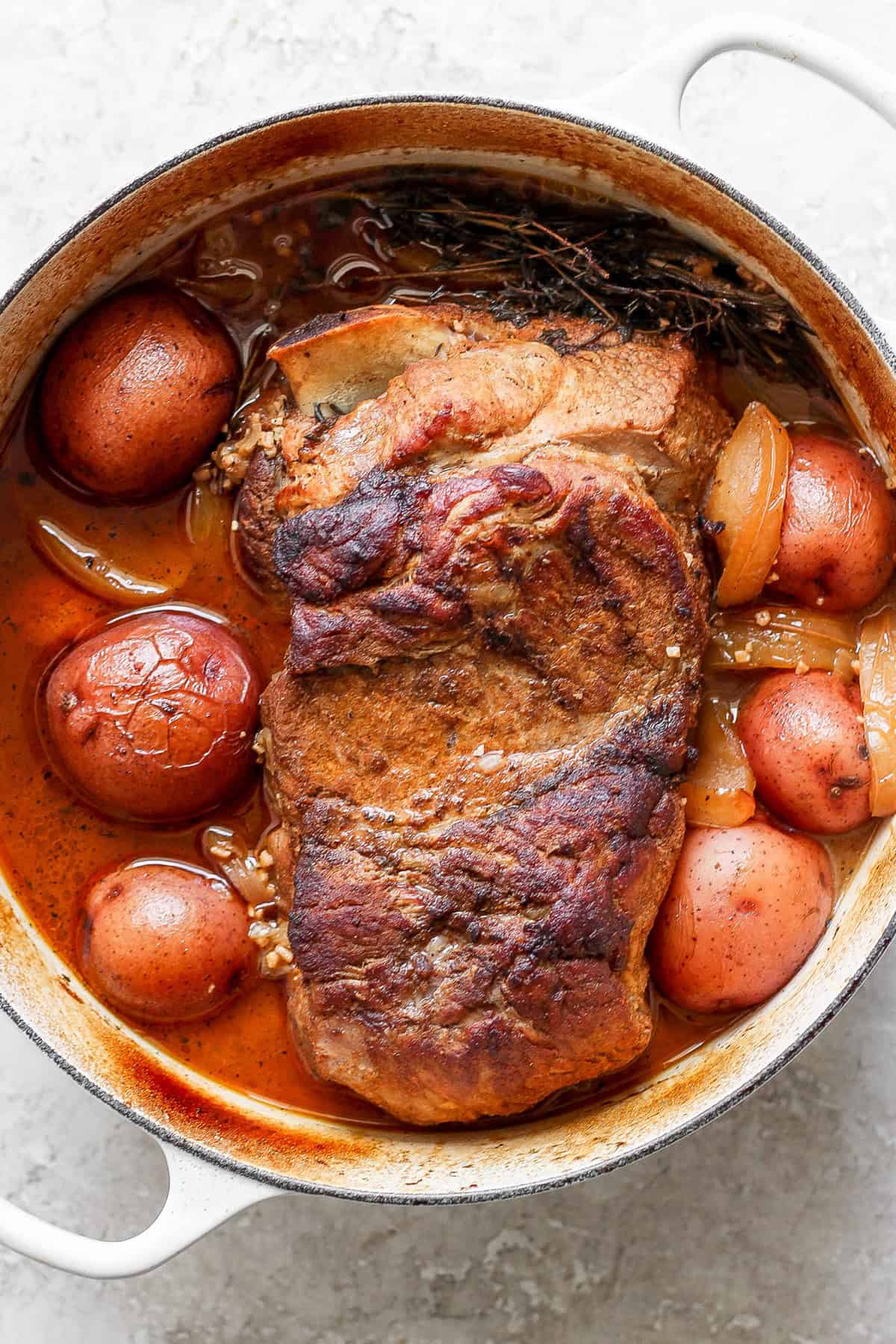 A cooked pot roast with potatoes and onions in a white enameled cast iron pot.