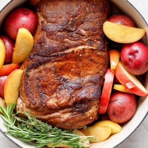 A cooked pork roast with a crispy exterior, surrounded by red potatoes, apple slices, and sprigs of rosemary in a white cast iron pot.