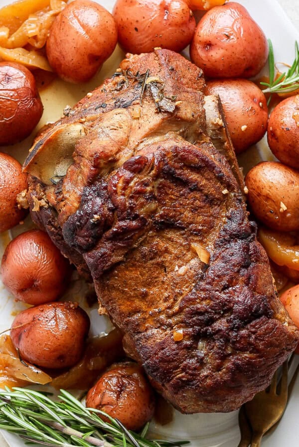 An oven-roasted pork roast surrounded by red potatoes and garnished with rosemary on a white platter.