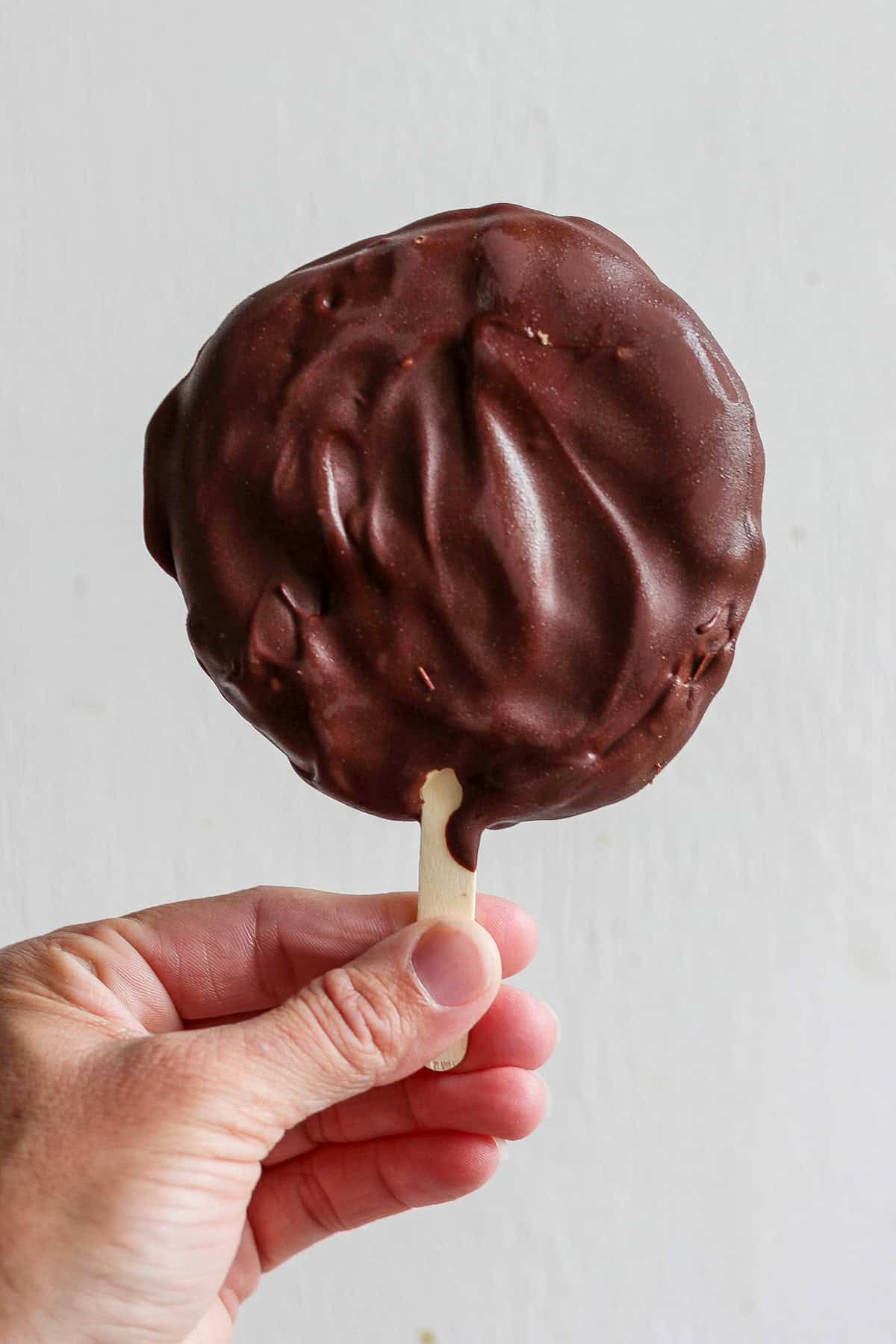 A hand holding a chocolate-covered ice cream on a stick against a plain background.