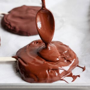 A spoon drizzles melted chocolate onto a round treat on a parchment-lined tray with a popsicle stick.