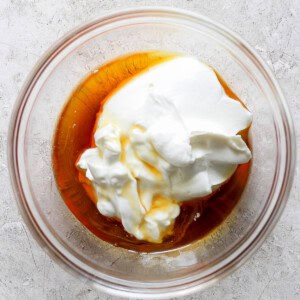 A glass bowl containing Greek yogurt and honey on a textured, light-colored surface.