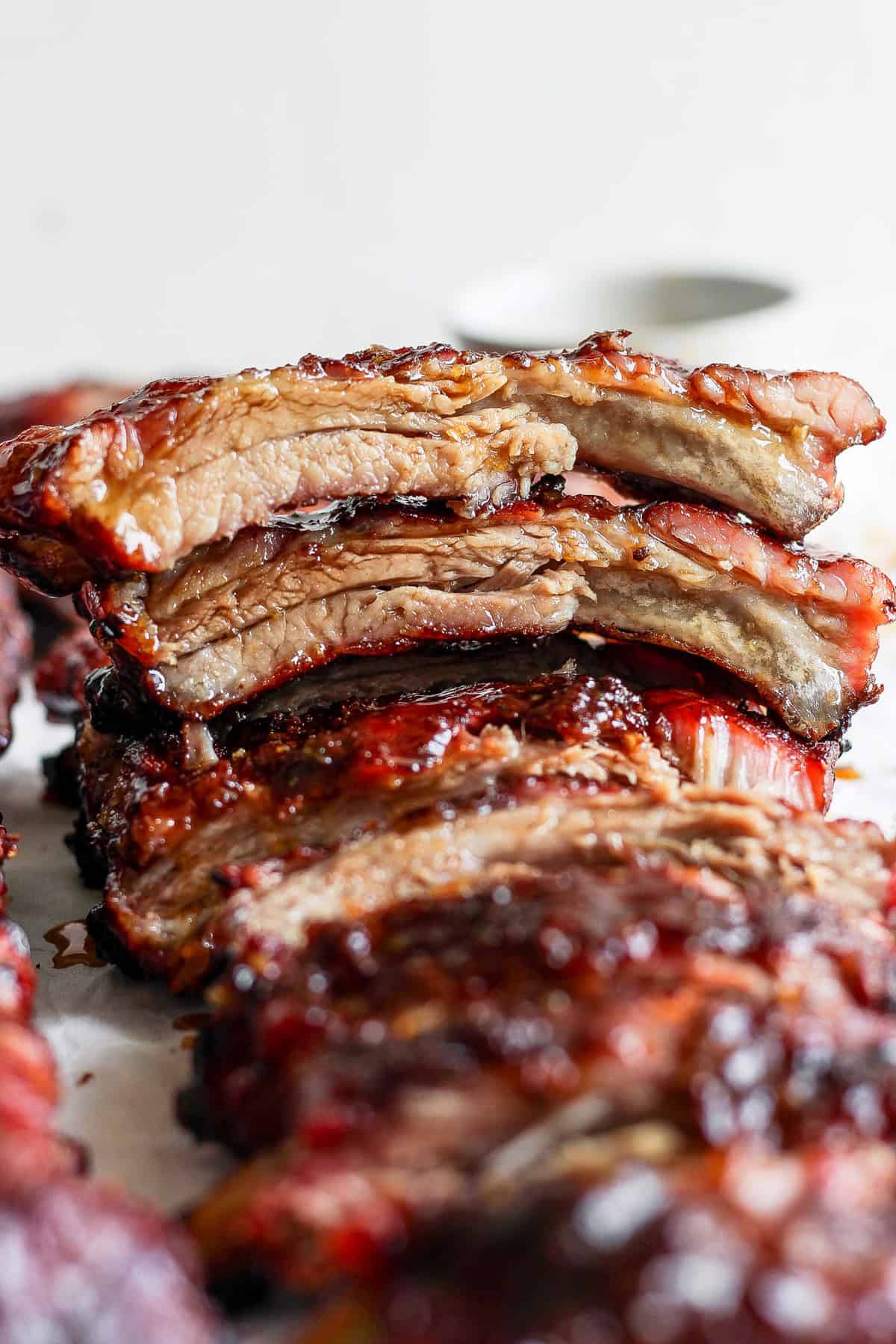 Close-up of a stack of glazed, grilled ribs, with the top piece cut to show a juicy, tender interior. The meat is richly caramelized with a glossy, charred exterior.