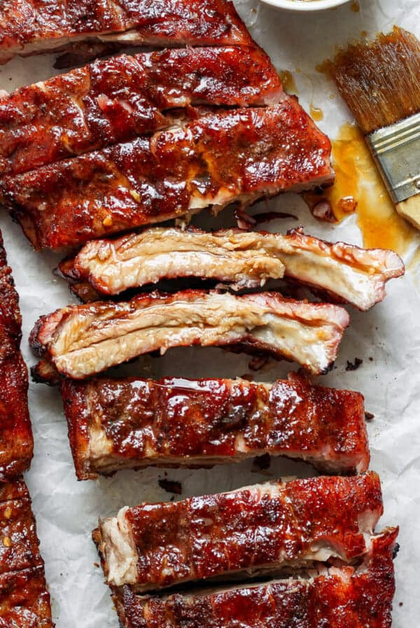 Close-up of cooked, glazed pork ribs on parchment paper with sauce and a basting brush beside them.