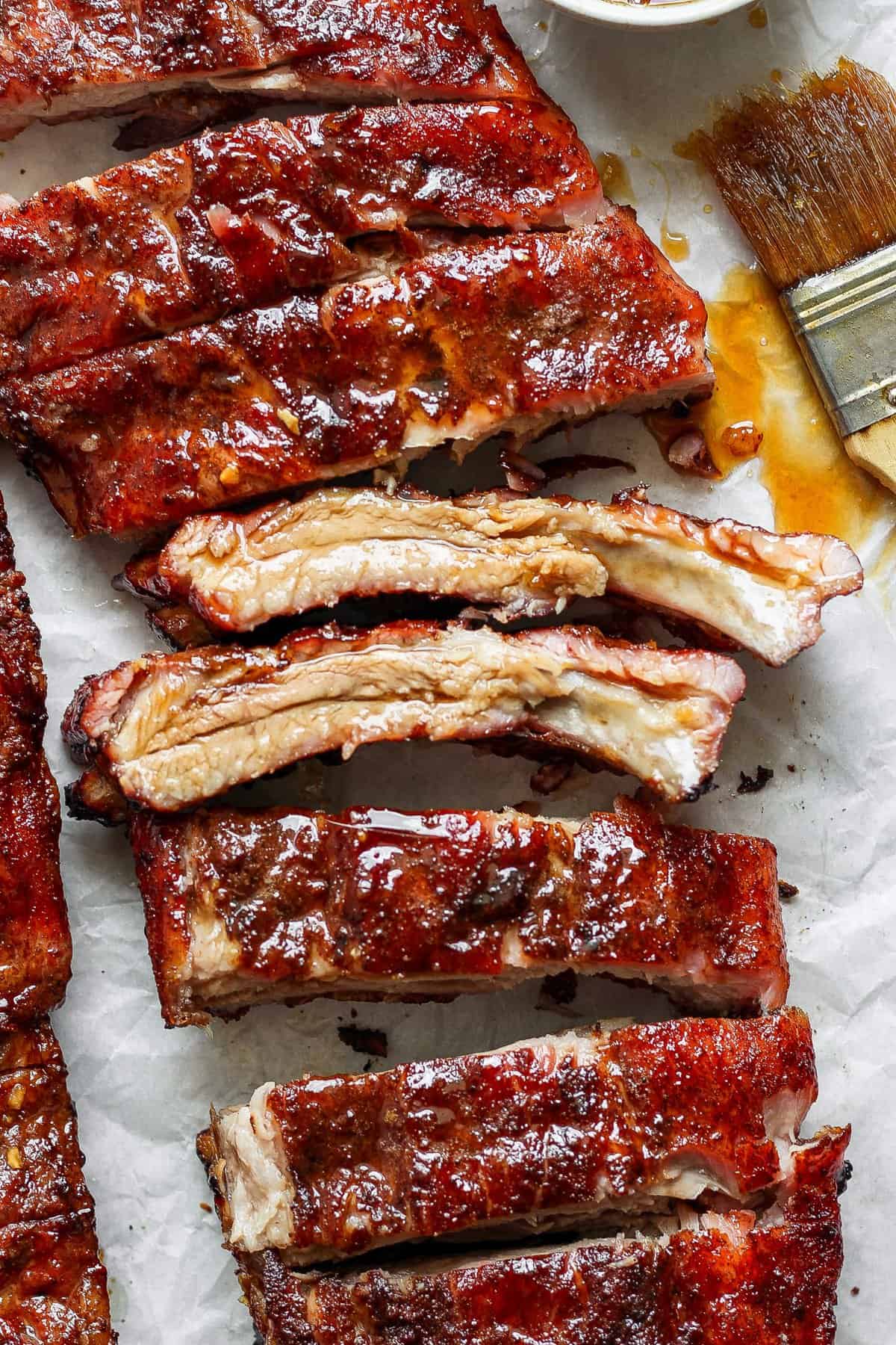 Close-up of cooked, glazed pork ribs on parchment paper with sauce and a basting brush beside them.