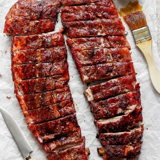 Two racks of barbecued ribs cut into portions, placed on parchment paper with a knife, a brush, and a bowl of sauce.