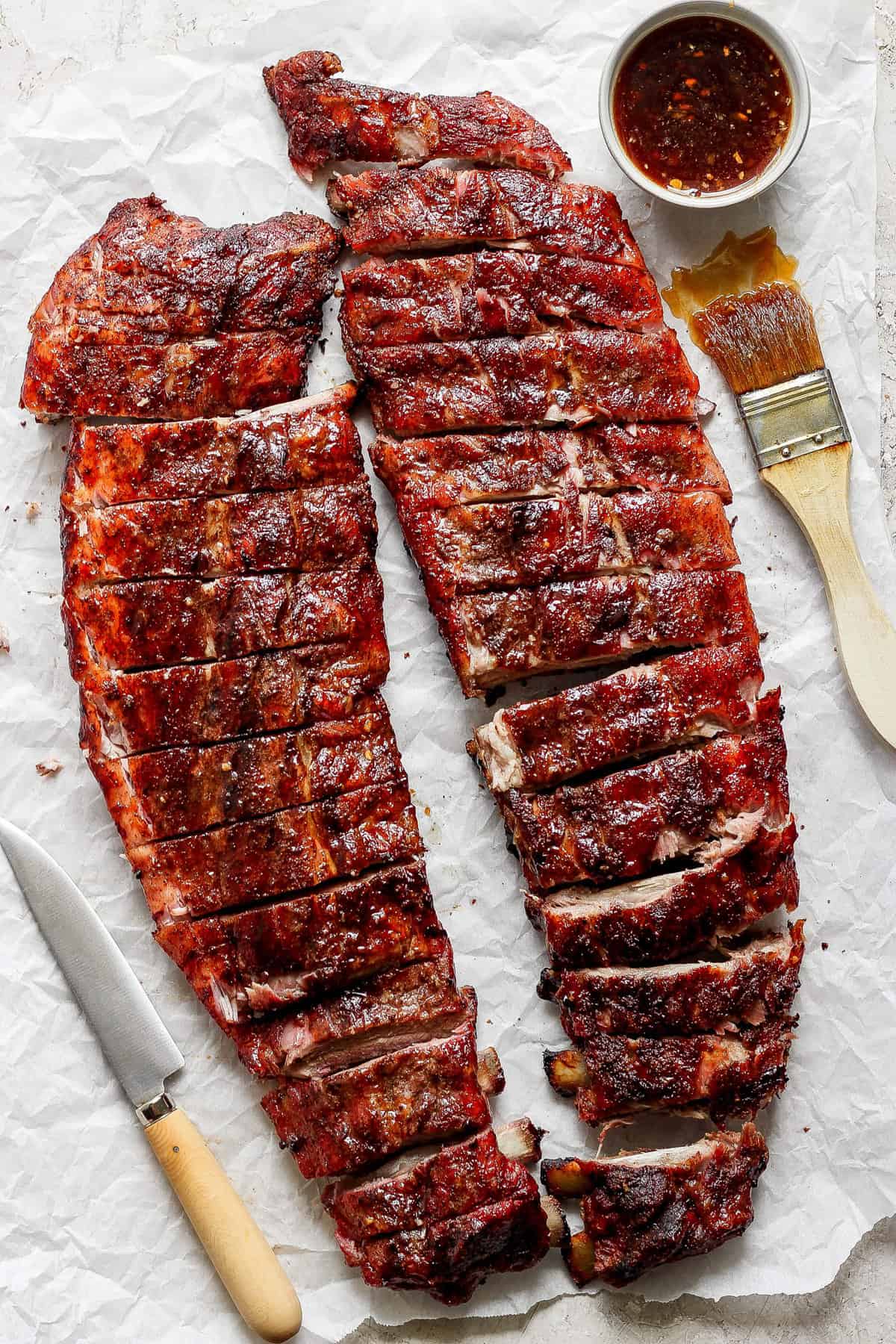 Two racks of barbecued ribs cut into portions, placed on parchment paper with a knife, a brush, and a bowl of sauce.