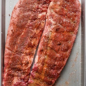 Two slabs of seasoned raw pork ribs are placed on a large rectangular baking sheet, ready for cooking.