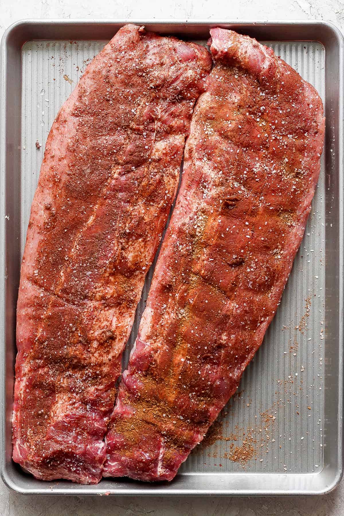Two slabs of seasoned raw pork ribs are placed on a large rectangular baking sheet, ready for cooking.