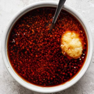 A white bowl filled with a reddish liquid and finely chopped ingredients, with a spoon and a dollop of minced garlic on top.