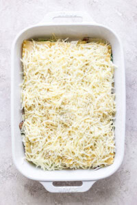 A rectangular white baking dish filled with unbaked lasagna, topped with a layer of shredded cheese, placed on a light-colored surface.