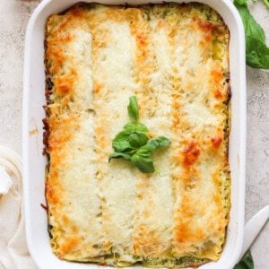 A baked lasagna in a white rectangular dish, topped with melted cheese and garnished with fresh basil leaves. A wooden pepper grinder and fresh basil are visible in the background.