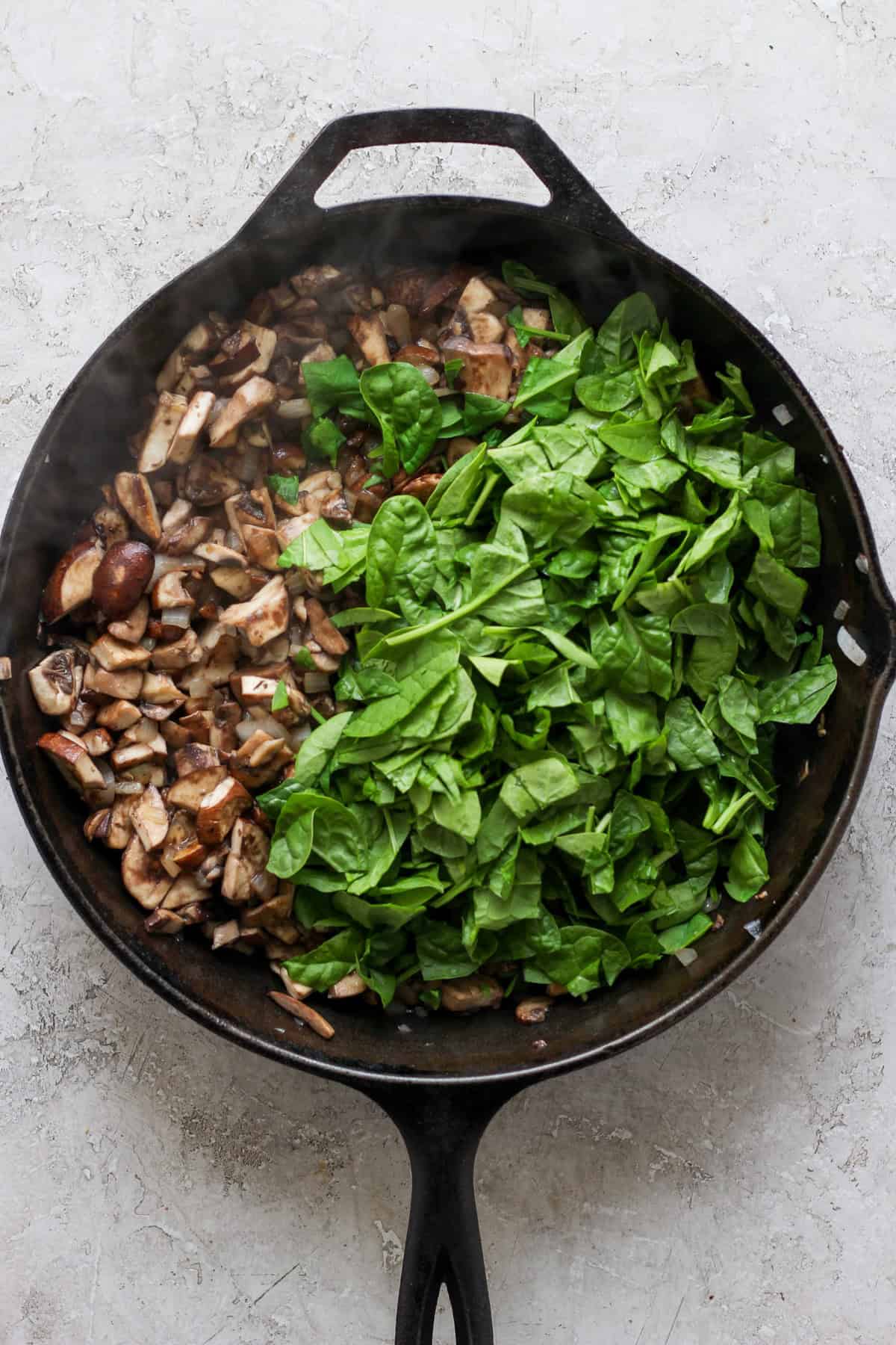 A cast-iron skillet filled with chopped mushrooms on one side and fresh spinach on the other, set on a light-colored surface.