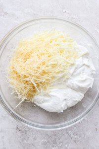 A glass bowl containing grated cheese and a mixture of white creamy ingredients on a light-colored surface.
