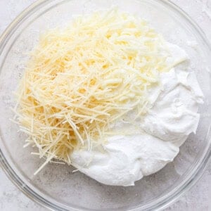 A glass bowl containing grated cheese and a mixture of white creamy ingredients on a light-colored surface.