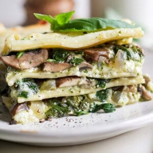 A slice of vegetable lasagna on a plate, featuring layers of pasta, mushrooms, spinach, ricotta, and topped with a basil leaf.