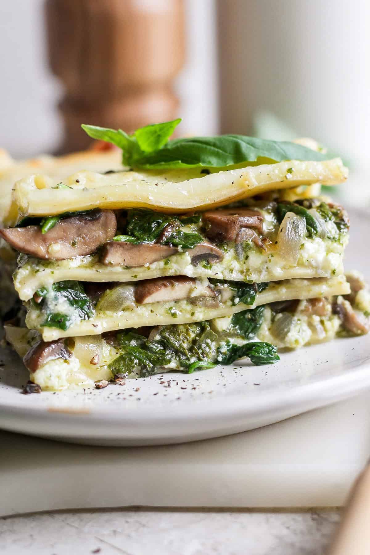 A slice of vegetable lasagna on a plate, featuring layers of pasta, mushrooms, spinach, ricotta, and topped with a basil leaf.