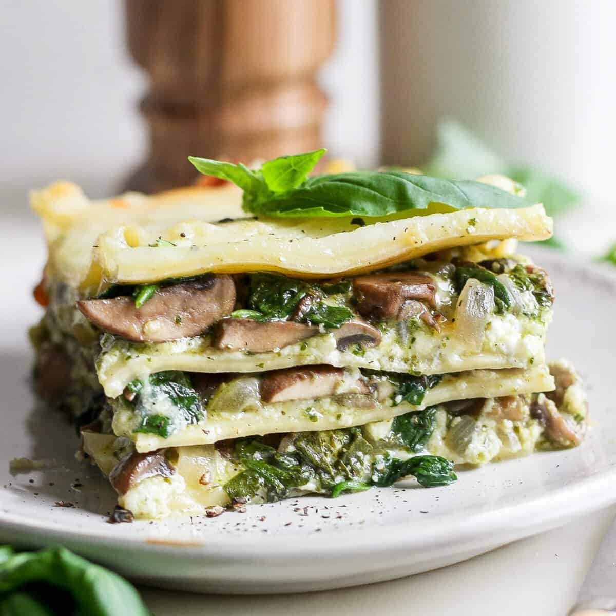 A slice of vegetable lasagna with visible layers of spinach, mushrooms, and cheese on a white plate, garnished with fresh basil leaves.