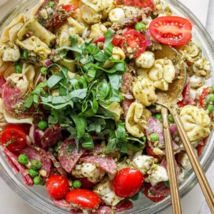 A bowl of tortellini pasta salad with cherry tomatoes, mozzarella, peas, salami, and greens, served with pesto and two spoons.