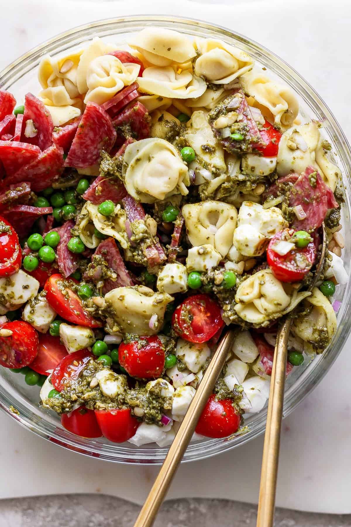 A bowl of pasta salad with tortellini, cherry tomatoes, peas, sliced meat, and pesto sauce, with two wooden utensils placed inside.