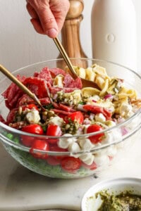 A glass bowl containing ingredients like cherry tomatoes, small mozzarella balls, salami slices, and pasta being mixed with spoons. A bottle and wooden salt grinder are in the background.