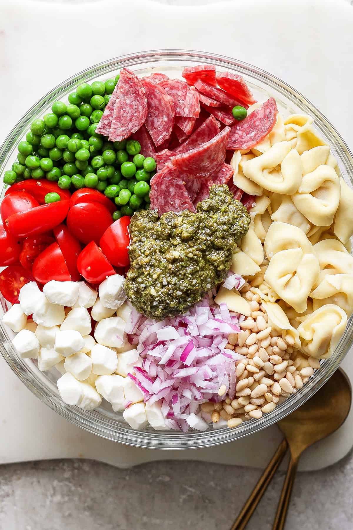 A glass bowl filled with peas, salami slices, cherry tomatoes, mozzarella balls, diced red onions, pine nuts, tortellini, and a dollop of pesto in the center. Two wooden utensils lie beside the bowl.