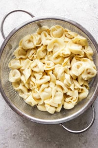 A metal colander filled with cooked cheese tortellini on a light-colored countertop.