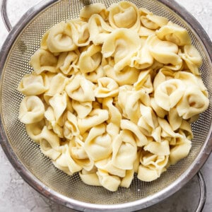 A metal colander filled with cooked cheese tortellini on a light-colored countertop.