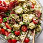 A close-up of a pasta salad with tortellini, cherry tomatoes, peas, mozzarella balls, salami, and green pesto sauce, served in a glass bowl with metal utensils.