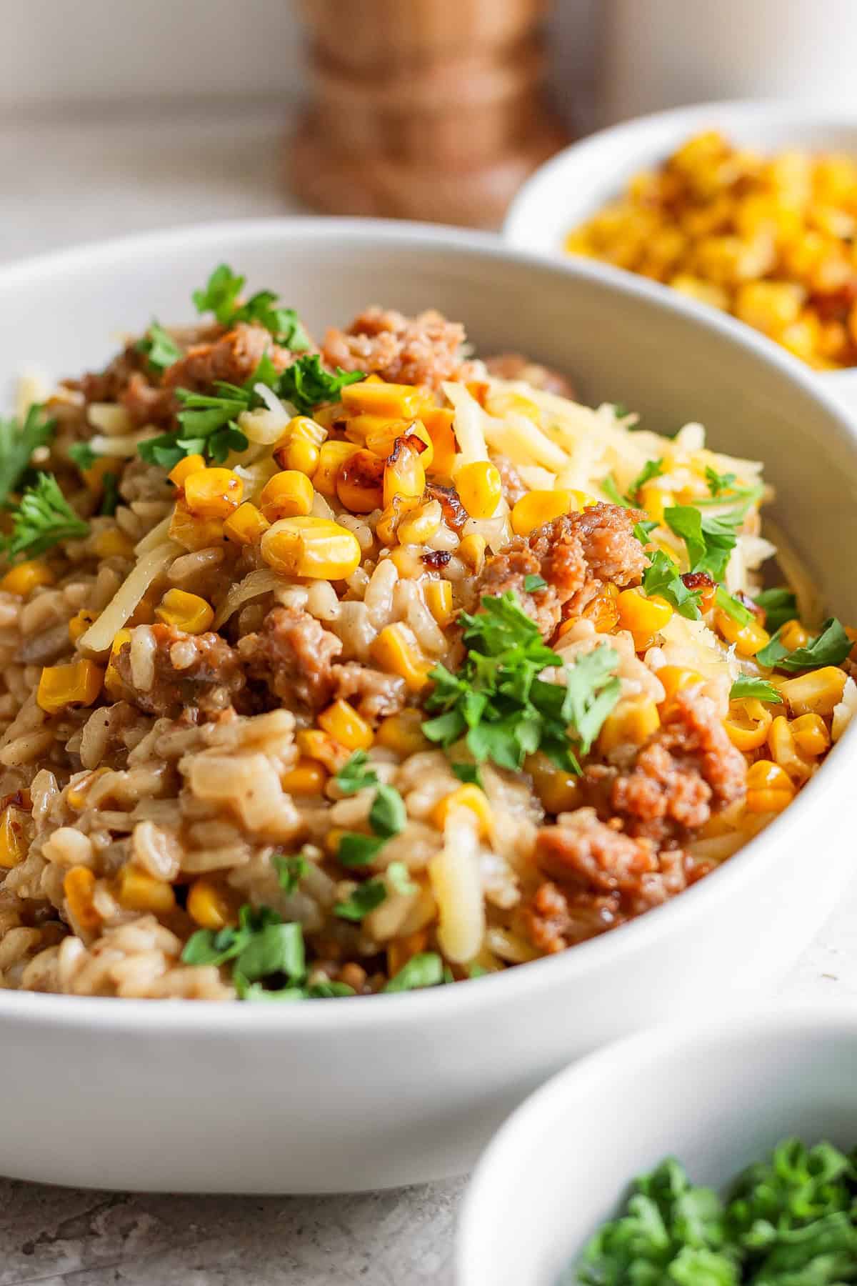 A bowl of risotto topped with ground meat, corn, shredded cheese, and fresh parsley. Other food dishes are blurred in the background.