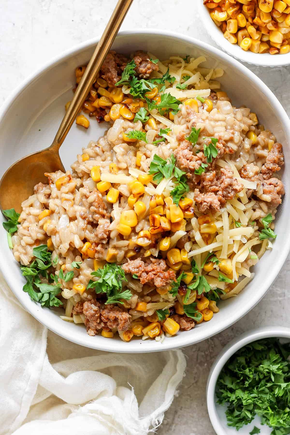 A bowl of barley risotto topped with corn, ground meat, shredded cheese, and garnished with fresh parsley. A gold spoon is placed on the side of the bowl. A small bowl of corn is in the background.