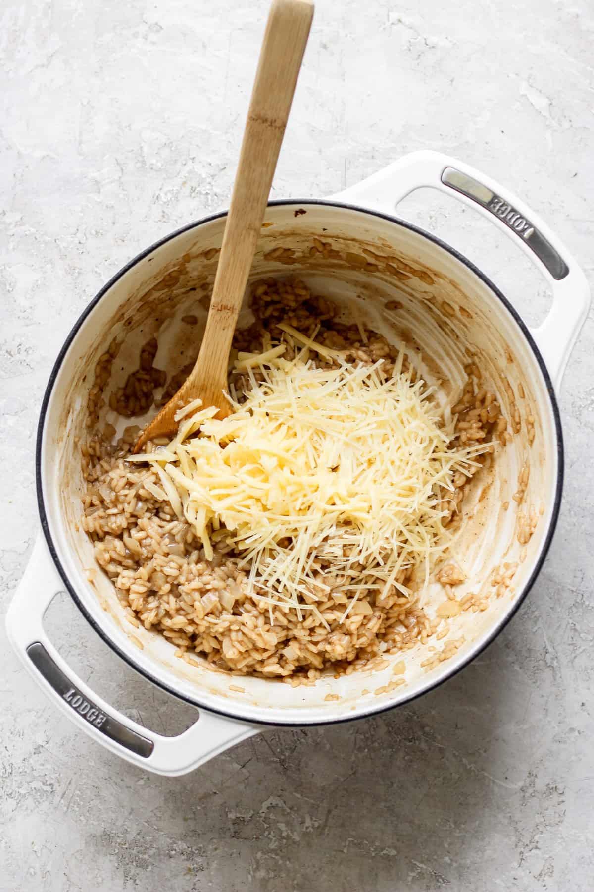 A wooden spoon rests in a white pot containing risotto, with grated cheese on top, ready to be stirred in.