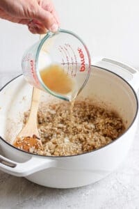 A person pours broth from a measuring cup into a white pot containing sautéed onions, stirring with a wooden spoon.