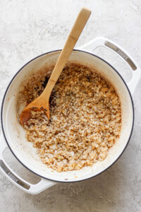 A white pot on a textured white surface contains sautéed onions and rice, being stirred with a wooden spoon.