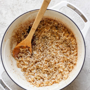 A white pot on a textured white surface contains sautéed onions and rice, being stirred with a wooden spoon.