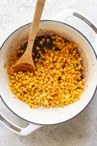 A wooden spoon rests in a white pot filled with seasoned, cooked corn kernels against a light gray background.