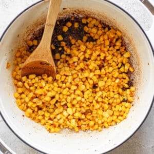 A wooden spoon rests in a white pot filled with seasoned, cooked corn kernels against a light gray background.