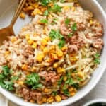 A bowl of risotto topped with ground meat, sweet corn, grated cheese, and chopped parsley with a gold-colored spoon on the side.