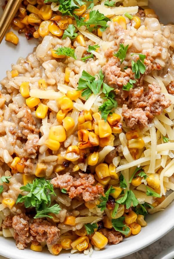 A bowl of risotto topped with ground meat, sweet corn, grated cheese, and chopped parsley with a gold-colored spoon on the side.