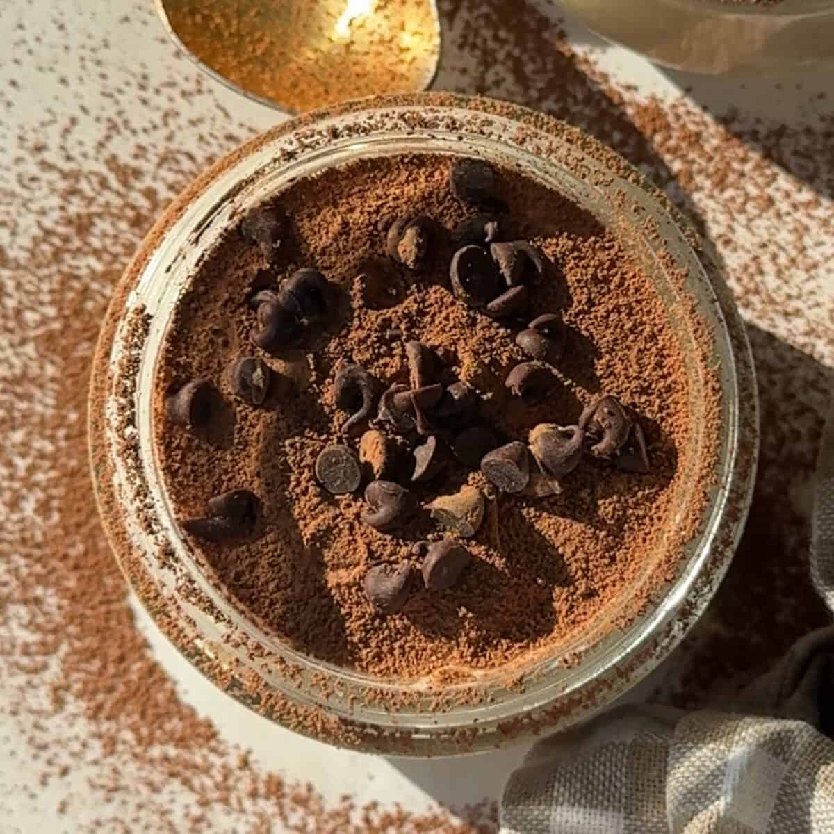 A jar filled with a powdered brown substance topped with chocolate chips hints at the delicious promise of blended overnight oats. A gold spoon is partially visible nearby, with more powder scattered on the surface.