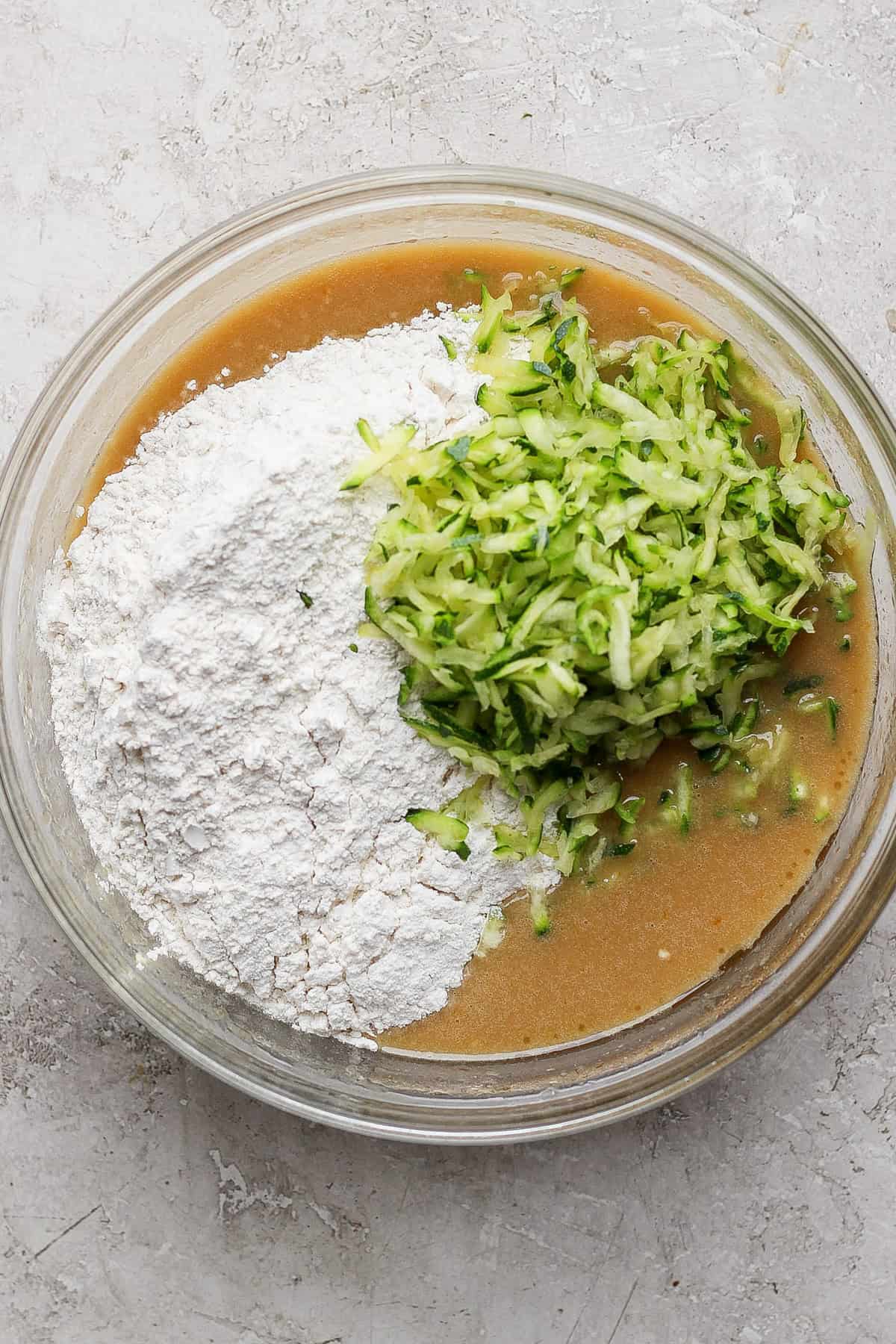 A glass bowl containing a baking mixture with flour, brown liquid batter, and grated zucchini on a light gray surface.