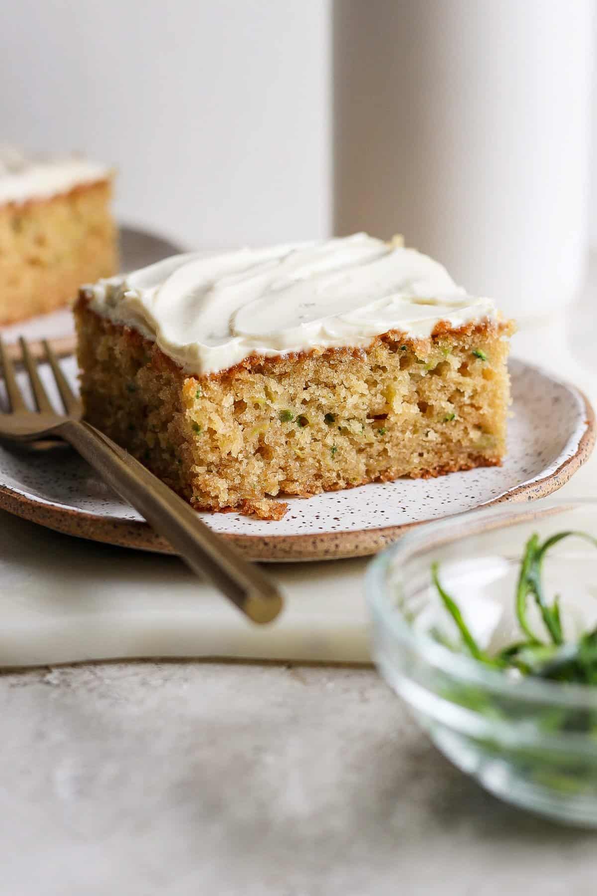 A slice of frosted zucchini cake on a plate with a fork beside it. A bowl with green garnish is in the foreground.