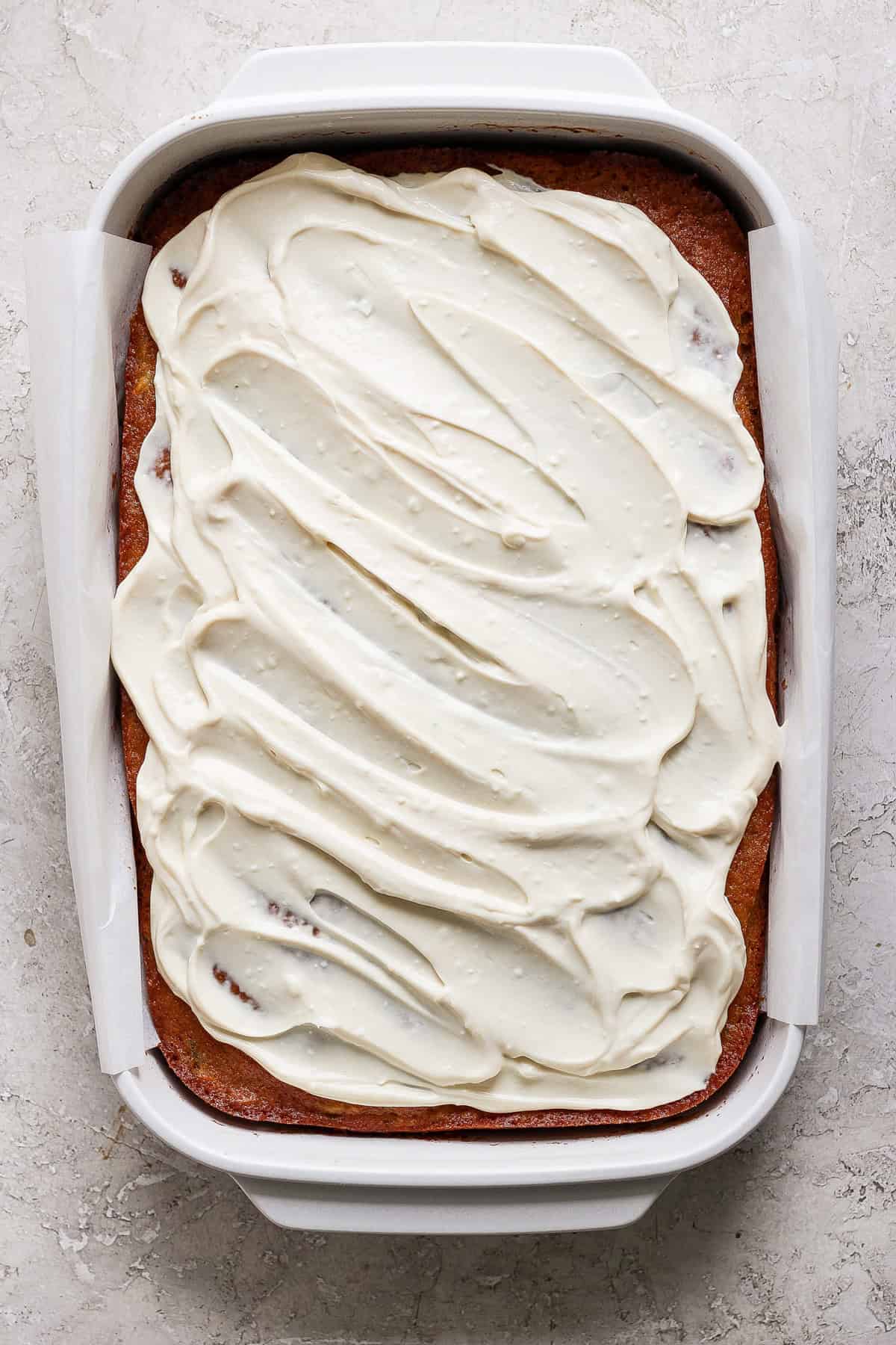 A rectangular cake with cream cheese frosting in a white baking dish, positioned on a light-colored surface.