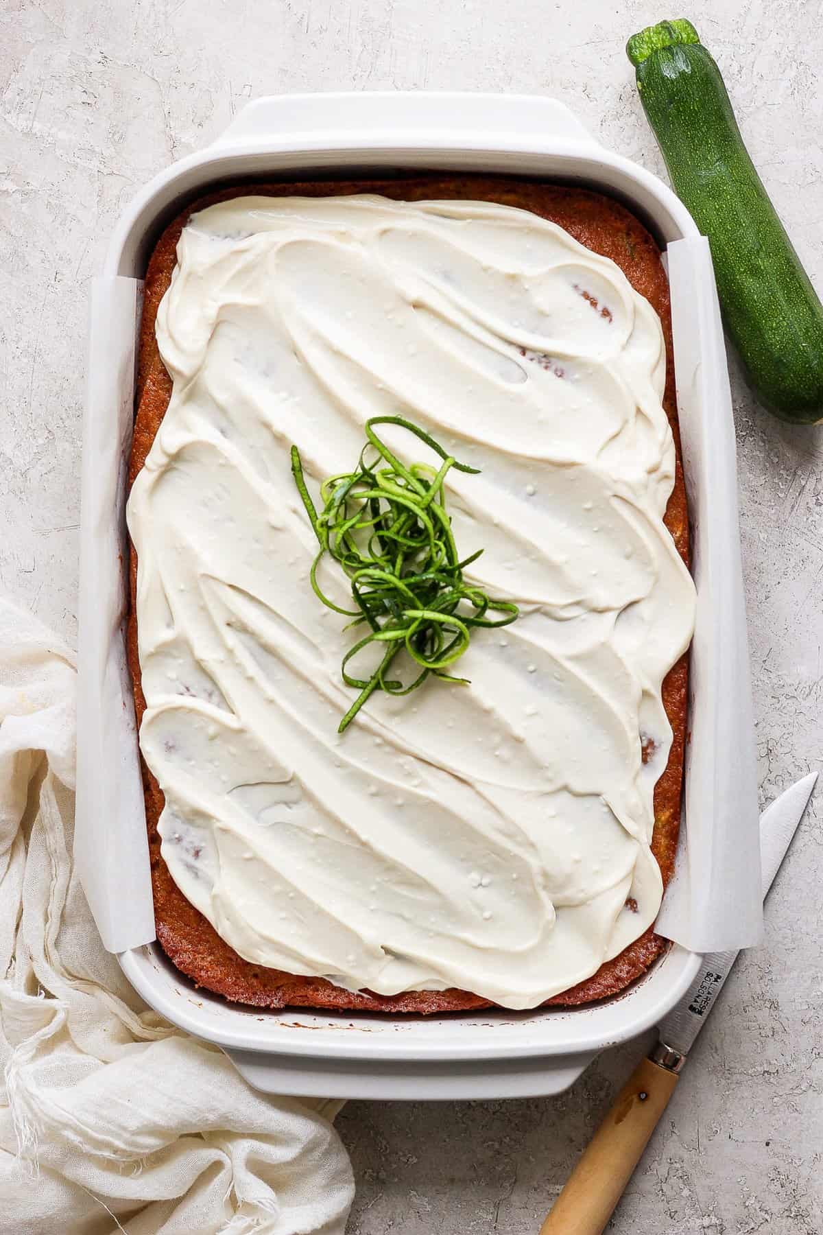 A frosted sheet cake garnished with spiralized zucchini sits in a rectangular baking dish. A knife, cloth, and a whole zucchini are placed beside the dish.