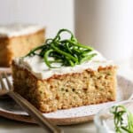 A slice of frosted zucchini cake topped with green zest strips, placed on a speckled plate with a fork beside it. Another slice is blurred in the background.