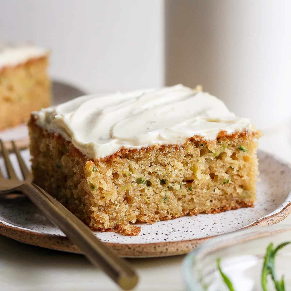 A slice of zucchini cake topped with cream cheese frosting on a speckled ceramic plate, with a fork beside it. Another slice is blurred in the background.